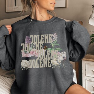 Op de afbeelding: Een donkergrijs sweatshirt met ronde hals en de herhaalde tekst "JOLENE" in een lichtbeige lettertype. De tekst is gelaagd met diverse botanische illustraties, waaronder paarse seringen, roze anjers, witte pioenrozen en groen gebladerte.