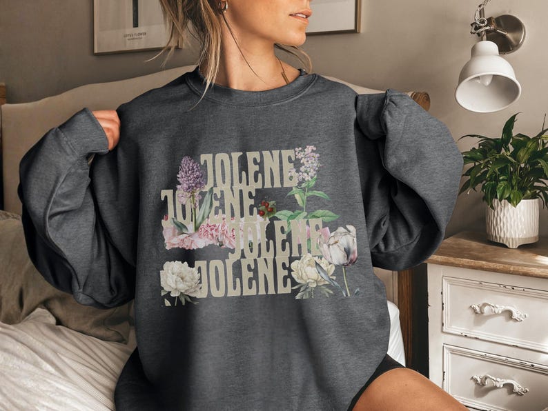 Op de afbeelding: Een donkergrijs sweatshirt met ronde hals en de herhaalde tekst "JOLENE" in een lichtbeige lettertype. De tekst is gelaagd met diverse botanische illustraties, waaronder paarse seringen, roze anjers, witte pioenrozen en groen gebladerte.