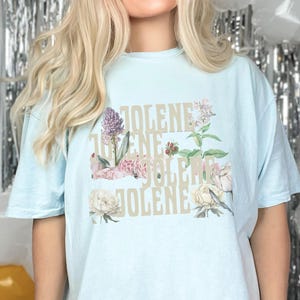 Comfort colors shirt in the color Chambray with the word Jolene in a staggered and stacked beige font written 4 times. There are wildflowers surrounding the words. Chambray is a light blue.