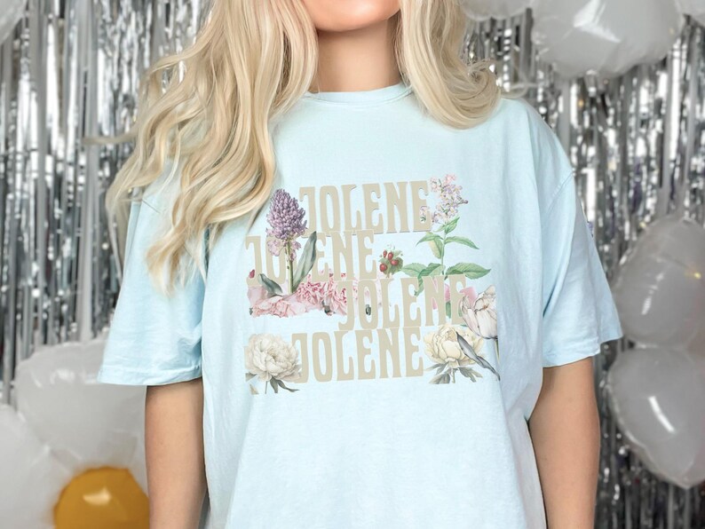 Comfort colors shirt in the color Chambray with the word Jolene in a staggered and stacked beige font written 4 times. There are wildflowers surrounding the words. Chambray is a light blue.