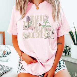 Op de afbeelding: Een lichtroze T-shirt met korte mouwen en een grafische print van de naam JOLENE, herhaald in een gestapeld, beige bloklettertype, gelaagd met illustraties van diverse kleurrijke bloemen, waaronder paarse, witte en groene botanische elementen.