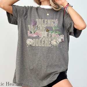 Op de afbeelding: Een donkergrijs t-shirt met korte mouwen en de herhaalde tekst JOLENE in een gestapeld, lichtgekleurd lettertype. Het ontwerp is versierd met diverse bloemenillustraties, waaronder paarse, roze en witte bloemen en groen blad.