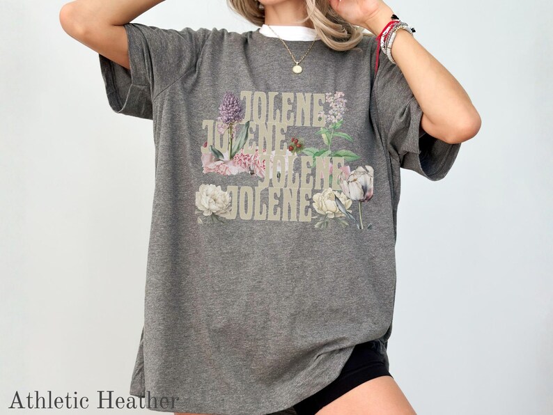 Op de afbeelding: Een donkergrijs t-shirt met korte mouwen en de herhaalde tekst JOLENE in een gestapeld, lichtgekleurd lettertype. Het ontwerp is versierd met diverse bloemenillustraties, waaronder paarse, roze en witte bloemen en groen blad.