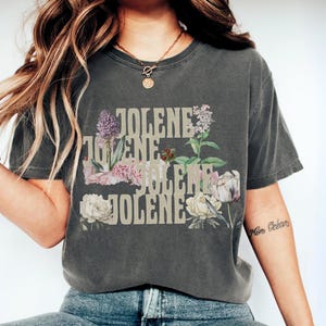 Op de afbeelding: Een antracietgrijs t-shirt met korte mouwen waarop de naam JOLENE vier keer onder elkaar is gedrukt in een lichtbeige bloklettertype. De tekst is gecombineerd met botanische illustraties van bloemen, waaronder witte pioenrozen, paarse seringen en groen blad.
