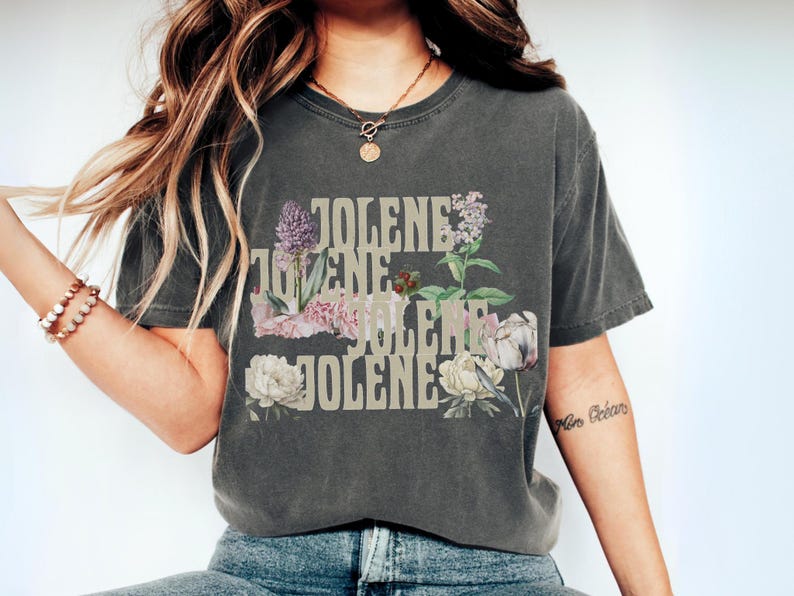 Op de afbeelding: Een antracietgrijs t-shirt met korte mouwen waarop de naam JOLENE vier keer onder elkaar is gedrukt in een lichtbeige bloklettertype. De tekst is gecombineerd met botanische illustraties van bloemen, waaronder witte pioenrozen, paarse seringen en groen blad.
