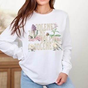 Op de afbeelding: Een wit shirt met lange mouwen met de tekst JOLENE herhaald in een gestapeld, lichtgekleurd lettertype. Het ontwerp is gelaagd met botanische illustraties van verschillende bloemen en groen in tinten roze, groen en cr&egrave;me.