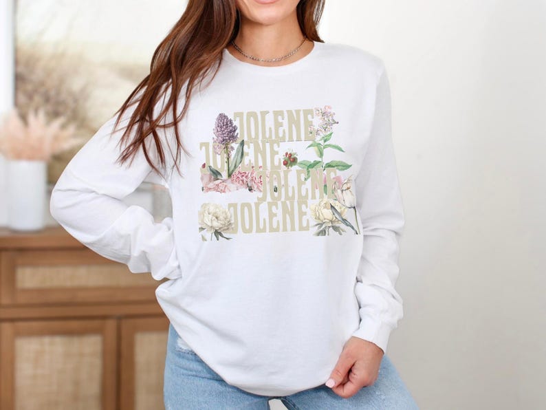 Op de afbeelding: Een wit shirt met lange mouwen met de tekst JOLENE herhaald in een gestapeld, lichtgekleurd lettertype. Het ontwerp is gelaagd met botanische illustraties van verschillende bloemen en groen in tinten roze, groen en cr&egrave;me.