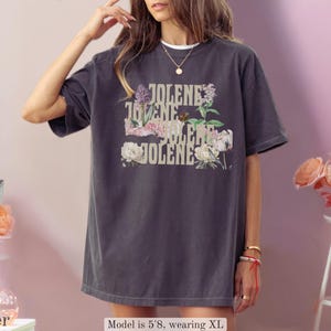 Op de afbeelding: Een antracietgrijs T-shirt met korte mouwen en de herhaalde tekst JOLENE in een beige lettertype, geplaatst over een bloemmotief met diverse bloemen in roze, witte en groene tinten. Het shirt heeft een ronde hals en een ruime, losse pasvorm.