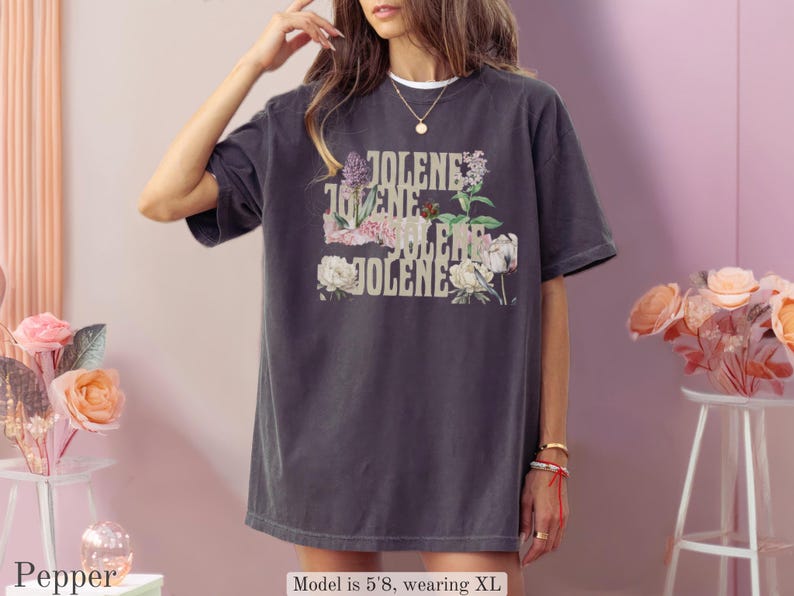 Op de afbeelding: Een antracietgrijs T-shirt met korte mouwen en de herhaalde tekst JOLENE in een beige lettertype, geplaatst over een bloemmotief met diverse bloemen in roze, witte en groene tinten. Het shirt heeft een ronde hals en een ruime, losse pasvorm.