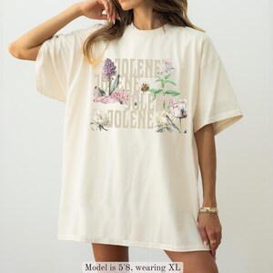 Op de afbeelding: Een ivoorkleurig t-shirt met de naam JOLENE in een herhalend verticaal patroon, gecombineerd met kleurrijke botanische illustraties van diverse bloemen en groene bladeren. Het shirt heeft een ronde hals en korte mouwen.