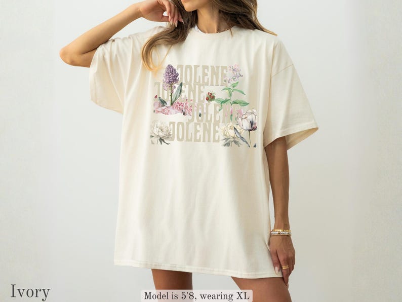 Op de afbeelding: Een ivoorkleurig t-shirt met de naam JOLENE in een herhalend verticaal patroon, gecombineerd met kleurrijke botanische illustraties van diverse bloemen en groene bladeren. Het shirt heeft een ronde hals en korte mouwen.
