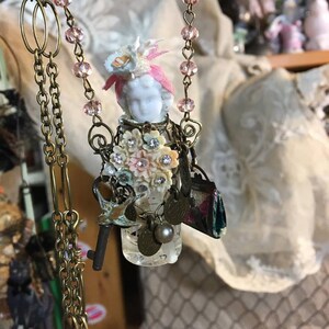 May include: A vintage-inspired necklace with a porcelain doll head, pink fabric flowers, and a variety of charms, including a key, a bird, and a small purse. The necklace is made with a gold chain and pink beads.