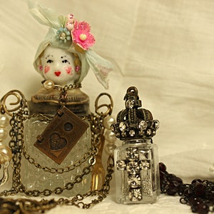 May include: Two vintage glass bottles with ornate metal tops and chains. One bottle has a porcelain doll head with a blue and white hat and pink flowers. The other bottle has a crown-shaped top with rhinestones. Both bottles are on a white fabric background.