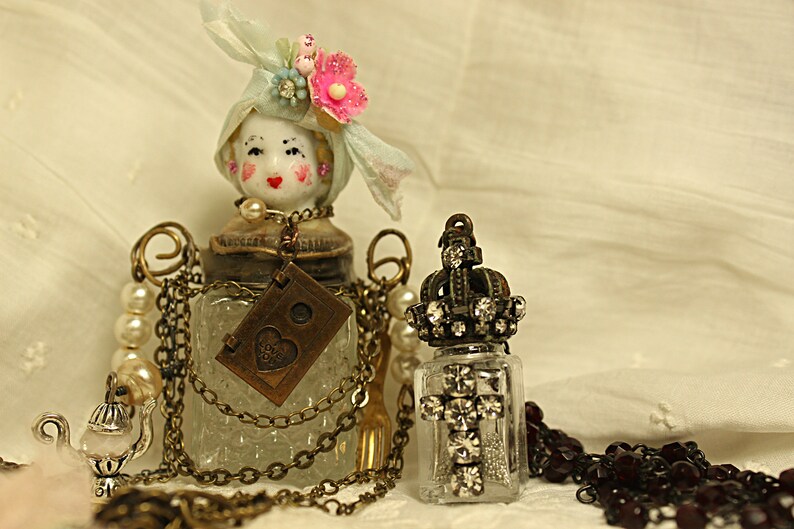 May include: Two vintage glass bottles with ornate metal tops and chains. One bottle has a porcelain doll head with a blue and white hat and pink flowers. The other bottle has a crown-shaped top with rhinestones. Both bottles are on a white fabric background.
