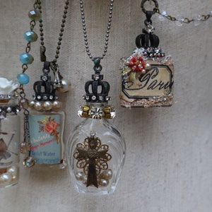 May include: Five glass bottle necklaces with different charms and embellishments. Each bottle has a crown-shaped stopper and is filled with various items, including dried flowers, pearls, and a cross. The necklaces are on a white mannequin.