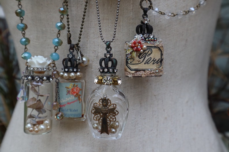 May include: Five glass bottle necklaces with different charms and embellishments. Each bottle has a crown-shaped stopper and is filled with various items, including dried flowers, pearls, and a cross. The necklaces are on a white mannequin.