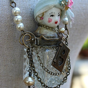 May include: A vintage-inspired glass bottle necklace with a porcelain doll head, a blue and white fabric head covering, and a small brass locket with a heart design. The necklace is adorned with pearl beads and brass chains.