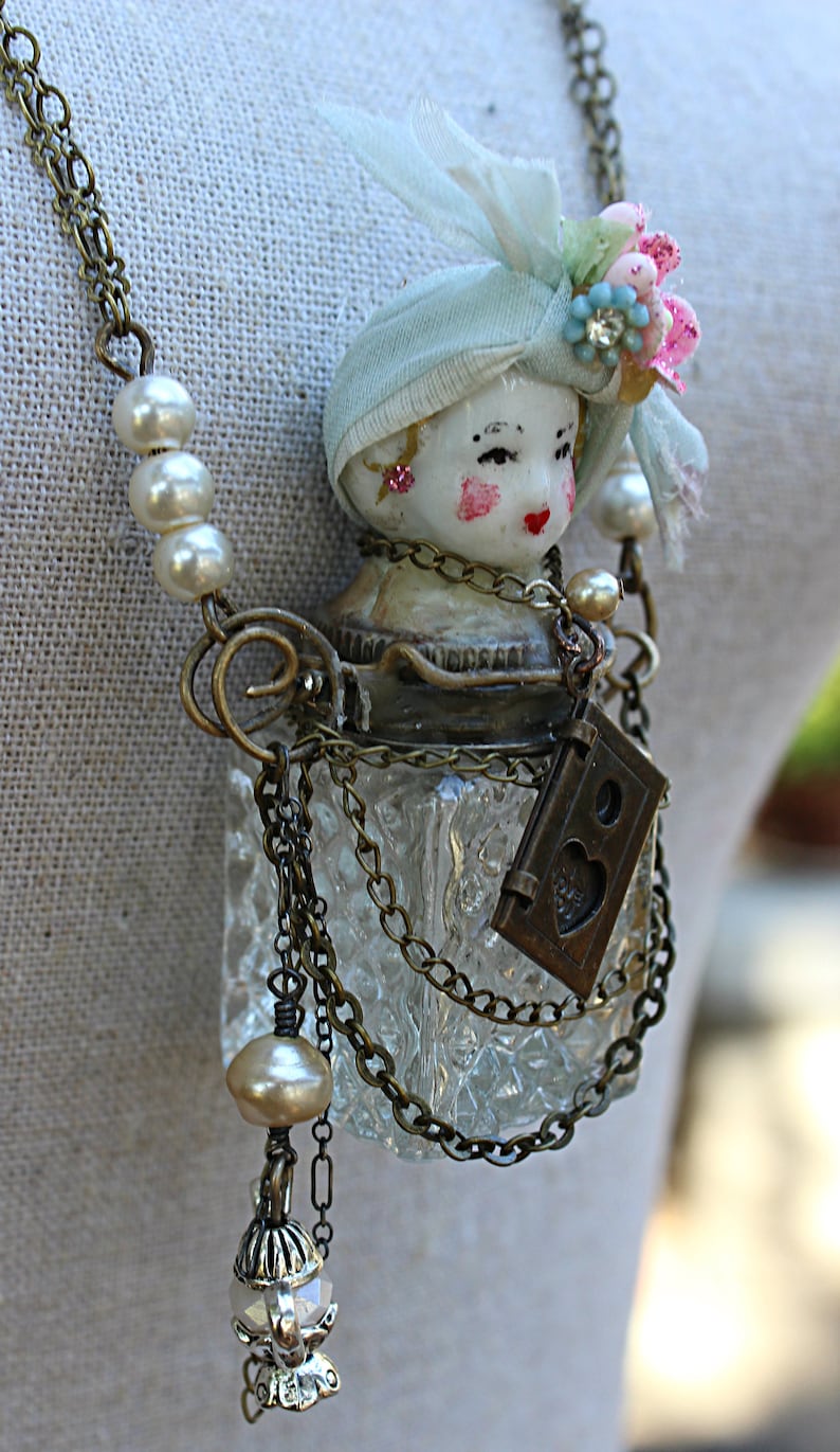 May include: A vintage-inspired glass bottle necklace with a porcelain doll head, a blue and white fabric head covering, and a small brass locket with a heart design. The necklace is adorned with pearl beads and brass chains.