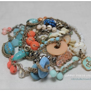 Peut inclure: Un collier &agrave; plusieurs rangs avec des perles bleues, corail, p&ecirc;che et blanches. Le collier est compos&eacute; d'un cordon crochet&eacute; et d'un charm avec un motif de couronne. Le texte "summer breeze crochet wrap" est visible.