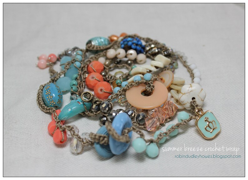 Peut inclure: Un collier &agrave; plusieurs rangs avec des perles bleues, corail, p&ecirc;che et blanches. Le collier est compos&eacute; d'un cordon crochet&eacute; et d'un charm avec un motif de couronne. Le texte "summer breeze crochet wrap" est visible.