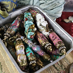 May include: A collection of colorful, fabric-wrapped beads with metallic accents and embellishments. The beads are in a silver tray and appear to be handmade.
