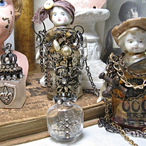 May include: Three decorative glass bottles with intricate embellishments and chains. The bottles are topped with porcelain doll heads and feature various charms and trinkets. The bottles are arranged on a vintage book with a brown leather cover.