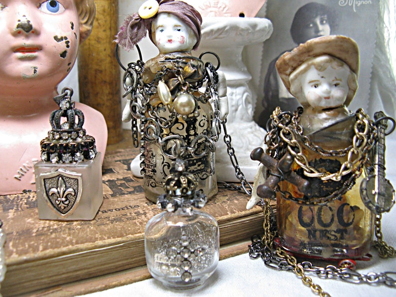 May include: Three decorative glass bottles with intricate embellishments and chains. The bottles are topped with porcelain doll heads and feature various charms and trinkets. The bottles are arranged on a vintage book with a brown leather cover.