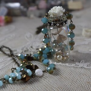 May include: A delicate necklace with a glass bottle pendant filled with white beads, seashells, and a white flower. The bottle is topped with a white flower and the necklace is made of a mix of blue and brown beads.