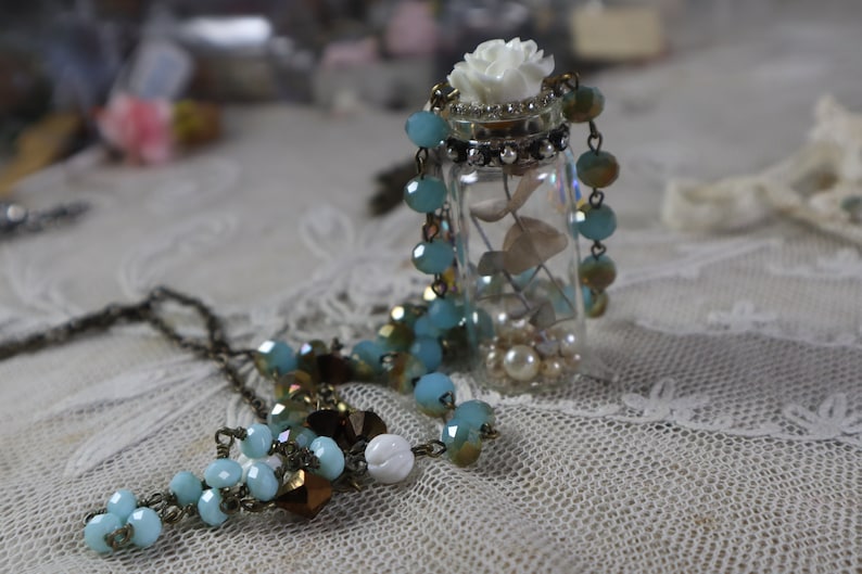 May include: A delicate necklace with a glass bottle pendant filled with white beads, seashells, and a white flower. The bottle is topped with a white flower and the necklace is made of a mix of blue and brown beads.