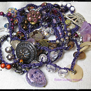 Peut inclure: Une collection de bijoux faits &agrave; la main avec une vari&eacute;t&eacute; de perles, de boutons et de breloques dans des tons de violet, de marron et de transparent. Les bijoux sont enfil&eacute;s sur un cordon violet et comprennent une breloque en m&eacute;tal avec l'inscription "Hand Made".