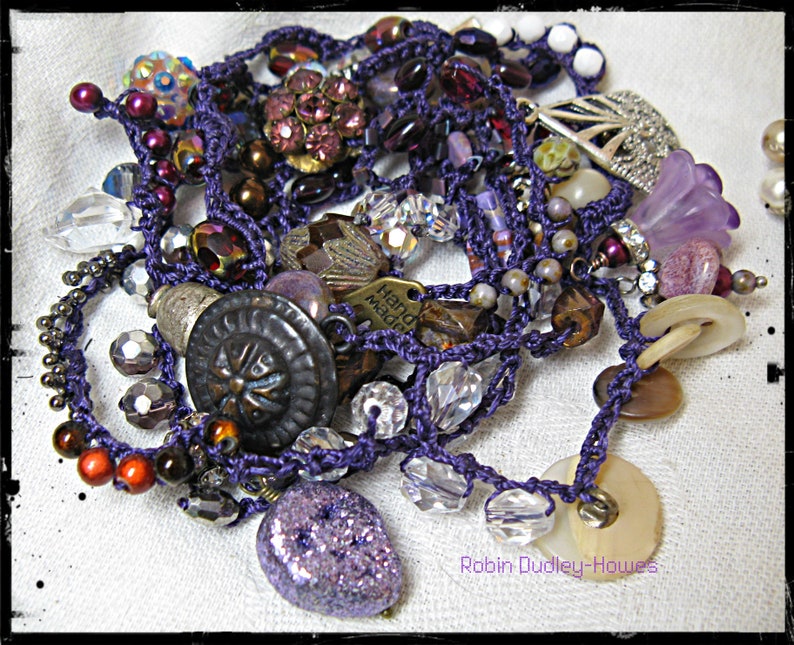 Peut inclure: Une collection de bijoux faits &agrave; la main avec une vari&eacute;t&eacute; de perles, de boutons et de breloques dans des tons de violet, de marron et de transparent. Les bijoux sont enfil&eacute;s sur un cordon violet et comprennent une breloque en m&eacute;tal avec l'inscription "Hand Made".