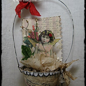 May include: A small paper pot with a white scalloped edge, filled with white tissue paper and a vintage image of a fairy with wings. The pot is decorated with a black and white printed design and the text "She's got a bit in her". A red ribbon and a wire handle are attached to the top of the pot.