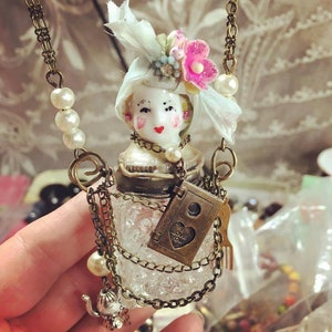May include: A vintage-inspired glass bottle necklace with a porcelain doll head, a delicate lace bow, and a charm that says "I love you". The necklace is adorned with multiple chains and pearl accents.