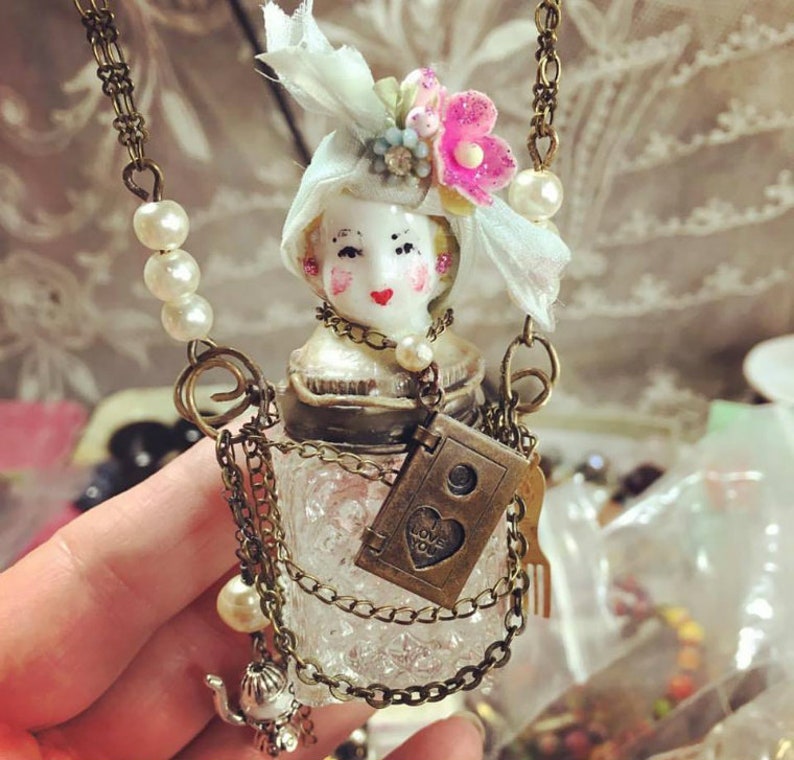 May include: A vintage-inspired glass bottle necklace with a porcelain doll head, a delicate lace bow, and a charm that says "I love you". The necklace is adorned with multiple chains and pearl accents.
