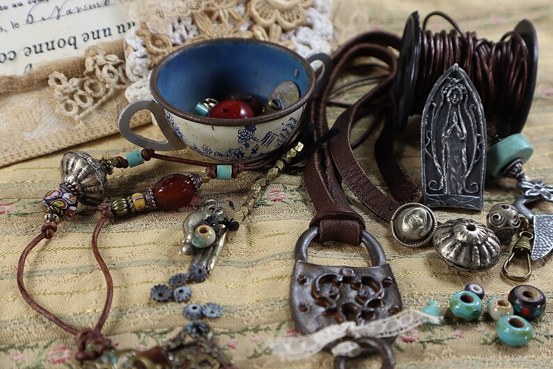 Peut inclure: Un assortiment de fournitures de fabrication de bijoux, comprenant des cordons en cuir, des perles, un cadenas d&eacute;coratif et une tasse &agrave; th&eacute; vintage remplie de perles. La palette de couleurs comprend le marron, l'argent, le bleu et le rouge.