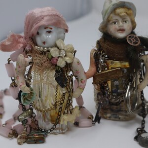 May include: Two porcelain dolls with painted faces and hats are decorated with vintage jewelry, beads, and fabric scraps. The dolls are attached to glass bottles with a variety of chains and charms.