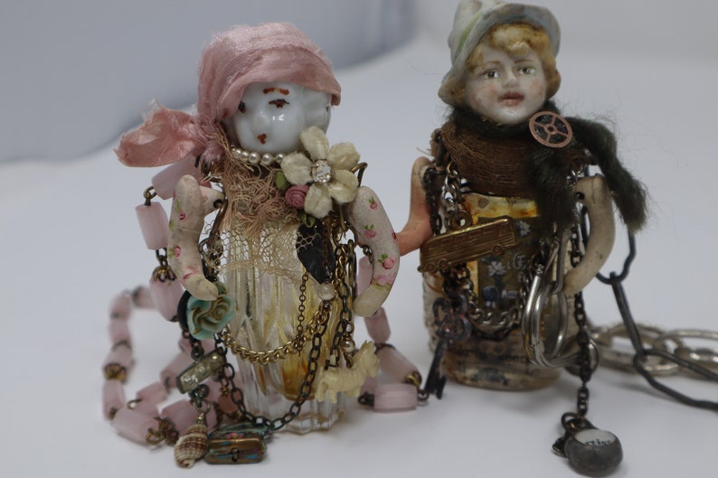May include: Two porcelain dolls with painted faces and hats are decorated with vintage jewelry, beads, and fabric scraps. The dolls are attached to glass bottles with a variety of chains and charms.