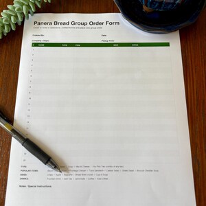 May include: A Panera Bread Group Order Form on a wooden surface, with a pen resting on it. The form has sections for company, items, and drinks. A small succulent plant in a blue ceramic pot is in the background.