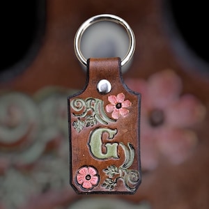 May include: A rectangular brown leather keychain featuring a silver metal ring and a silver rivet. The leather is embossed with a large green letter G, surrounded by intricate carved floral patterns with pink and green accents.