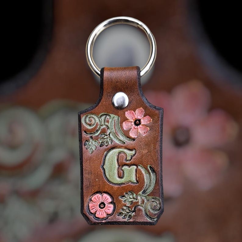 May include: A rectangular brown leather keychain featuring a silver metal ring and a silver rivet. The leather is embossed with a large green letter G, surrounded by intricate carved floral patterns with pink and green accents.