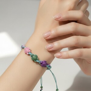 May include: A handmade bracelet featuring three turtle charms in pink, green, and purple. The turtles are strung on a blue cord with adjustable ties. The bracelet is shown on a wrist.