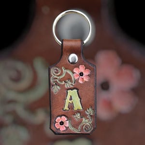 May include: A brown leather keychain with a gold-colored key ring. The keychain features the letter "A" in a light green color, along with pink flowers and decorative green leaves. The background is blurred.