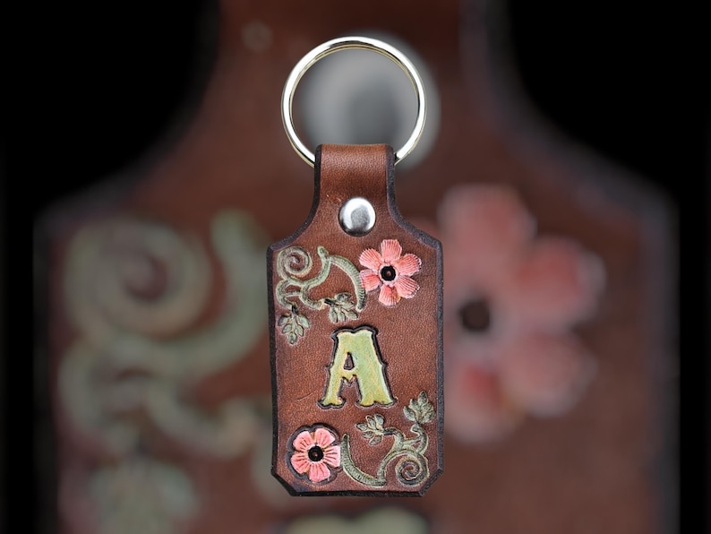 May include: A brown leather keychain with a gold-colored key ring. The keychain features the letter "A" in a light green color, along with pink flowers and decorative green leaves. The background is blurred.