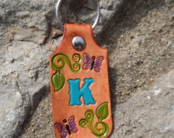May include: Leather keychain with a turquoise letter K, green vines, and purple butterflies. The keychain is attached to a silver key ring.