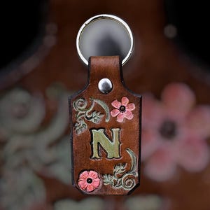 May include: A brown leather keychain with a silver ring. The keychain features the letter "N" in green, surrounded by pink flowers and green leaves. A silver rivet secures the ring to the leather.