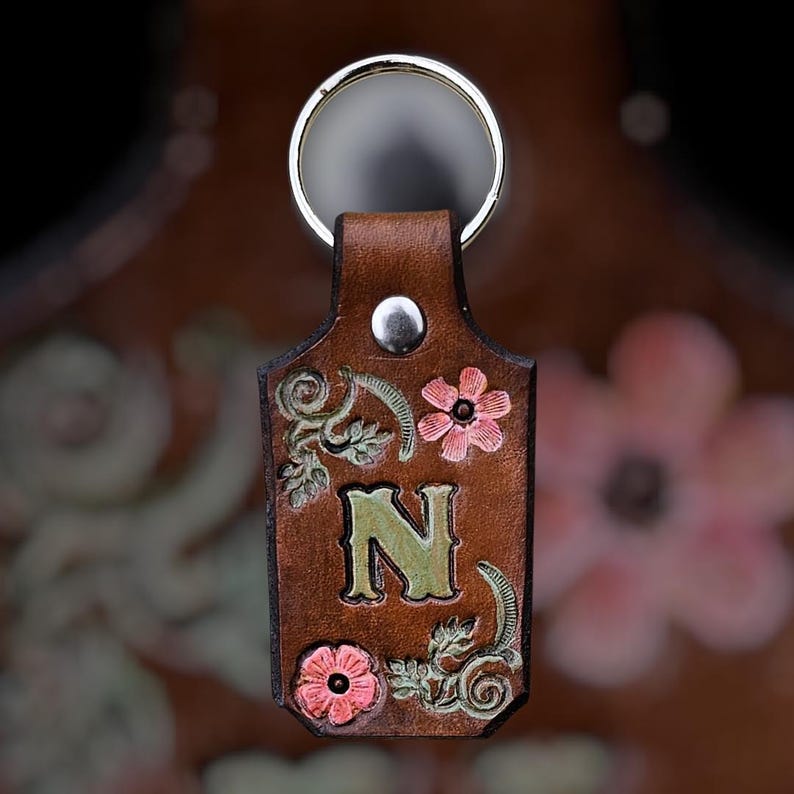 May include: A brown leather keychain with a silver ring. The keychain features the letter "N" in green, surrounded by pink flowers and green leaves. A silver rivet secures the ring to the leather.