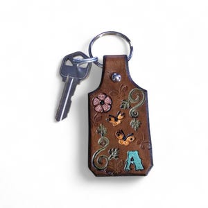 May include: A brown leather keychain with a silver key attached. The keychain is decorated with embossed flowers, butterflies, and the letter "A" in teal. The keychain is attached to a silver key ring.