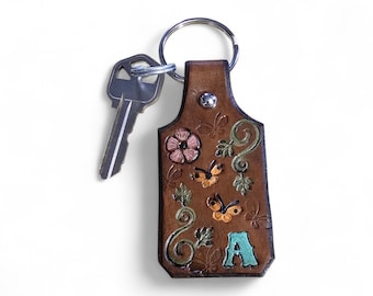 May include: A brown leather keychain with a silver key attached. The keychain is decorated with embossed flowers, butterflies, and the letter "A" in teal. The keychain is attached to a silver key ring.