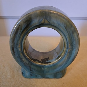 May include: A ceramic ring holder with a green and brown glaze. The ring holder has a circular opening in the center.