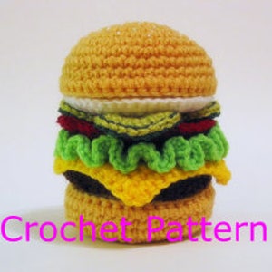 Może przedstawiać: Amigurumi burger zrobiony na szydełku z żółtą bułką, zieloną sałatą, czerwonym pomidorem, żółtym serem i brązowym kotletem.